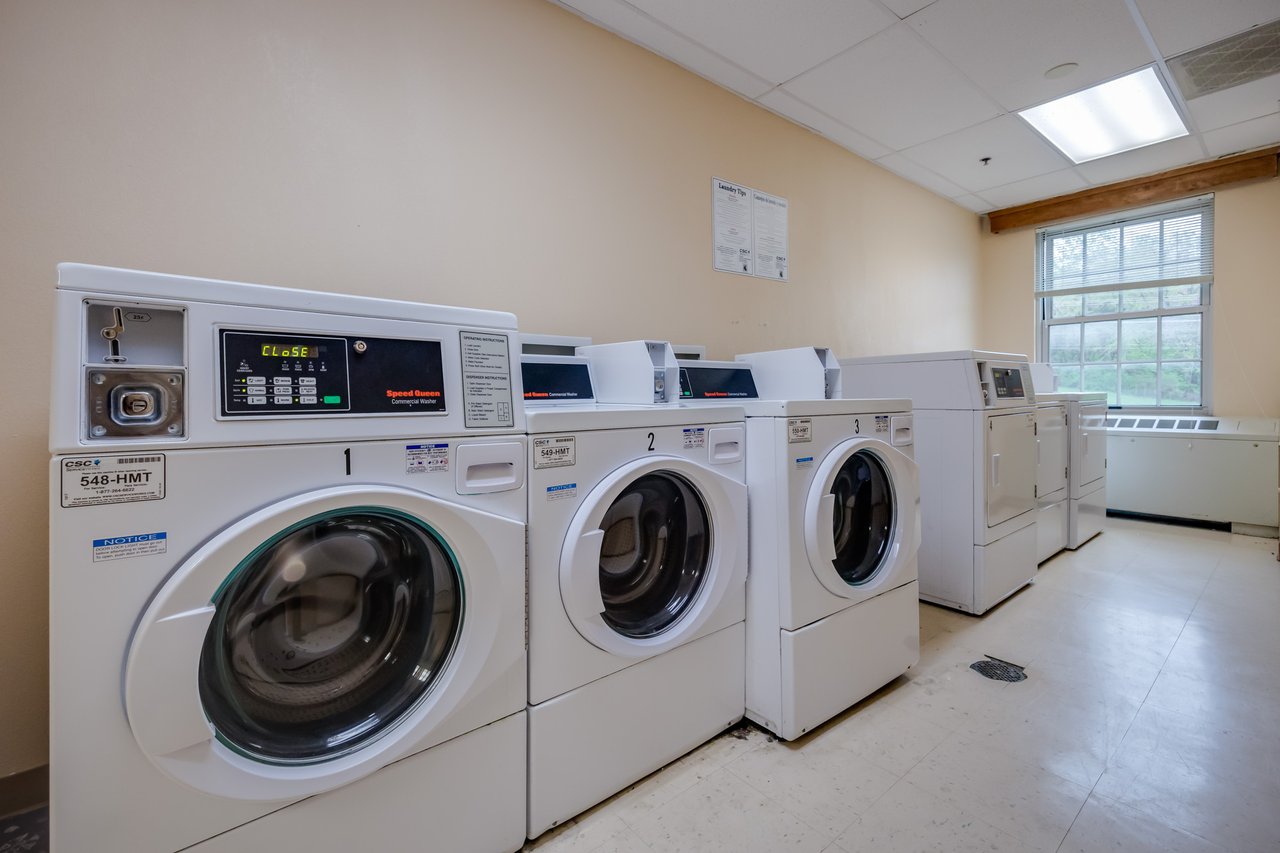 Washers and dryers are lined up on the wall of a laundry room with tan walls and a tile floor.