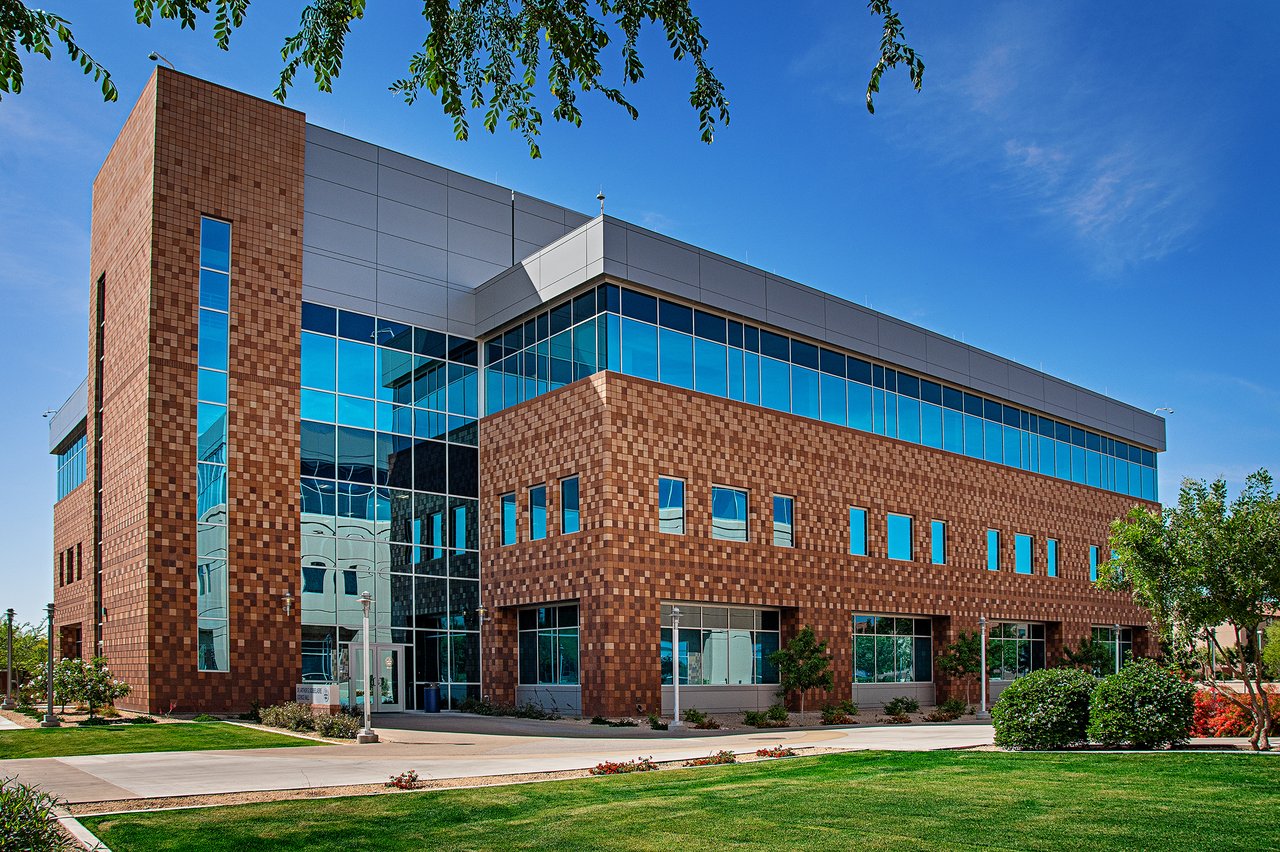 Modern building with a brown brick exterior and large glass windows, surrounded by greenery and a clear blue sky.