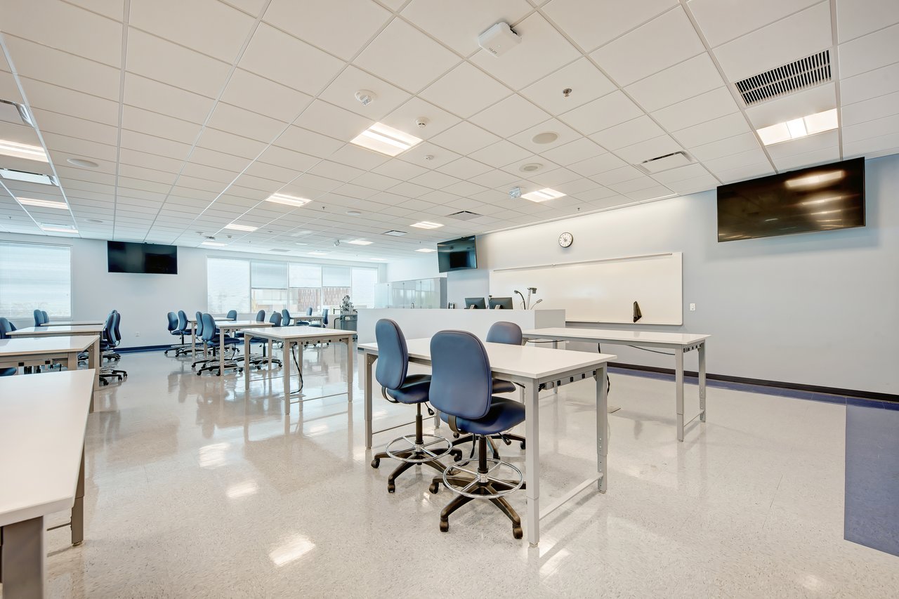 A modern classroom featuring rows of tables, blue chairs, large windows, and mounted screens.