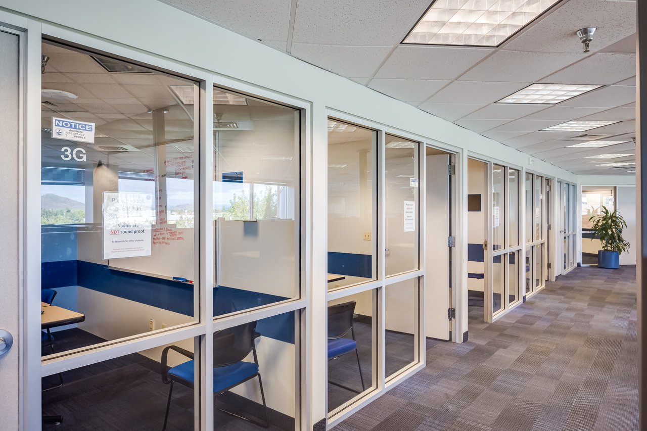 Modern office corridor with glass-walled meeting rooms and blue chairs, featuring natural light and a plant.