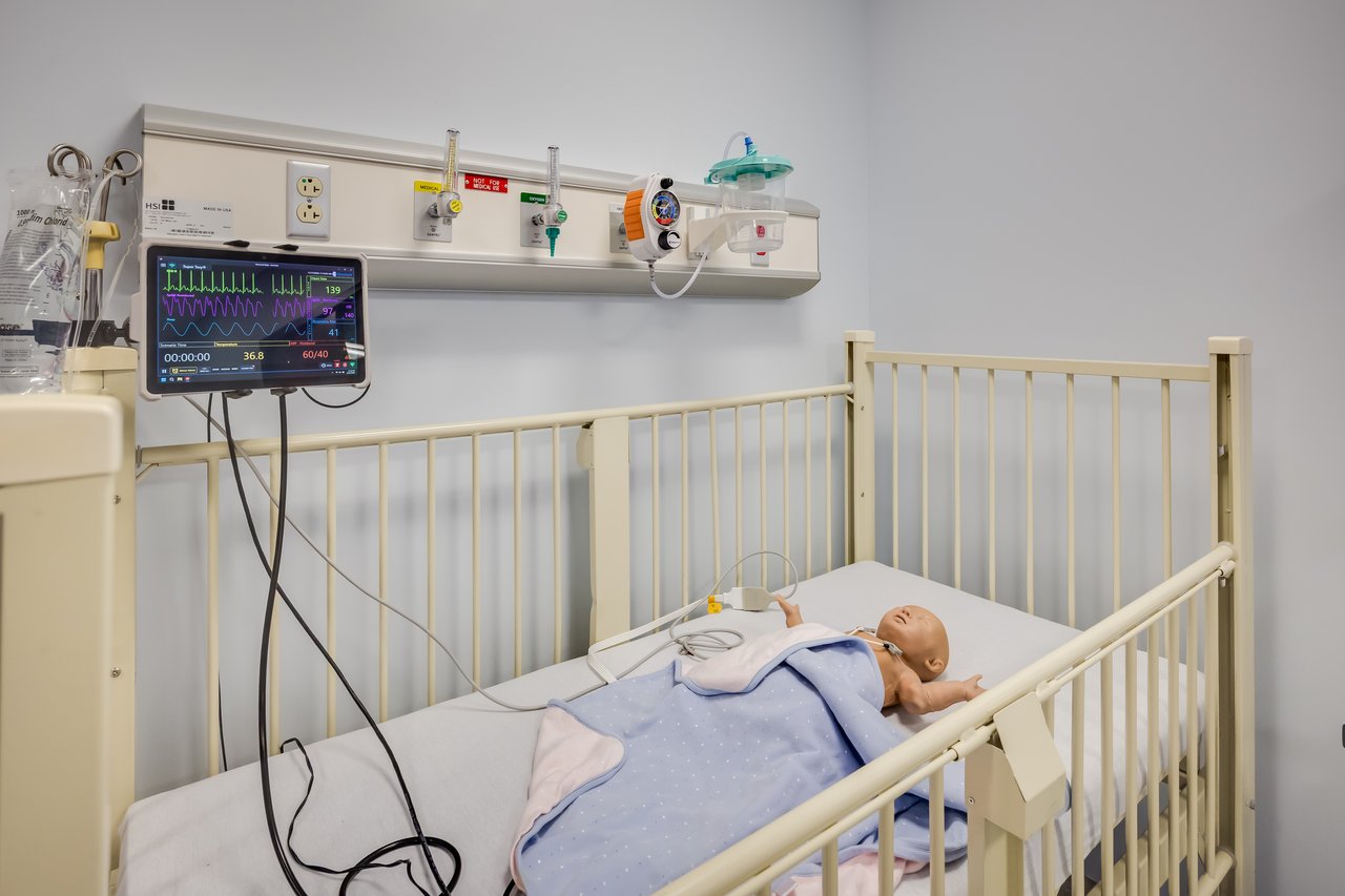 A medical cot with a baby mannequin, a monitor displaying vital signs, and medical equipment on the wall.