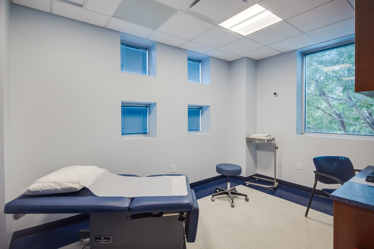 Medical exam room with a blue examination table, a stool, and large windows, creating a bright, inviting atmosphere.