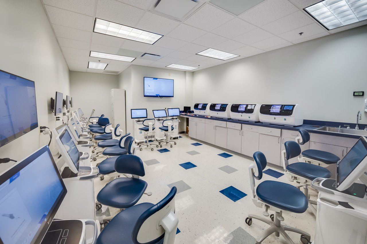 A modern laboratory with rows of computers and ergonomic chairs, featuring equipment along the wall and screens.