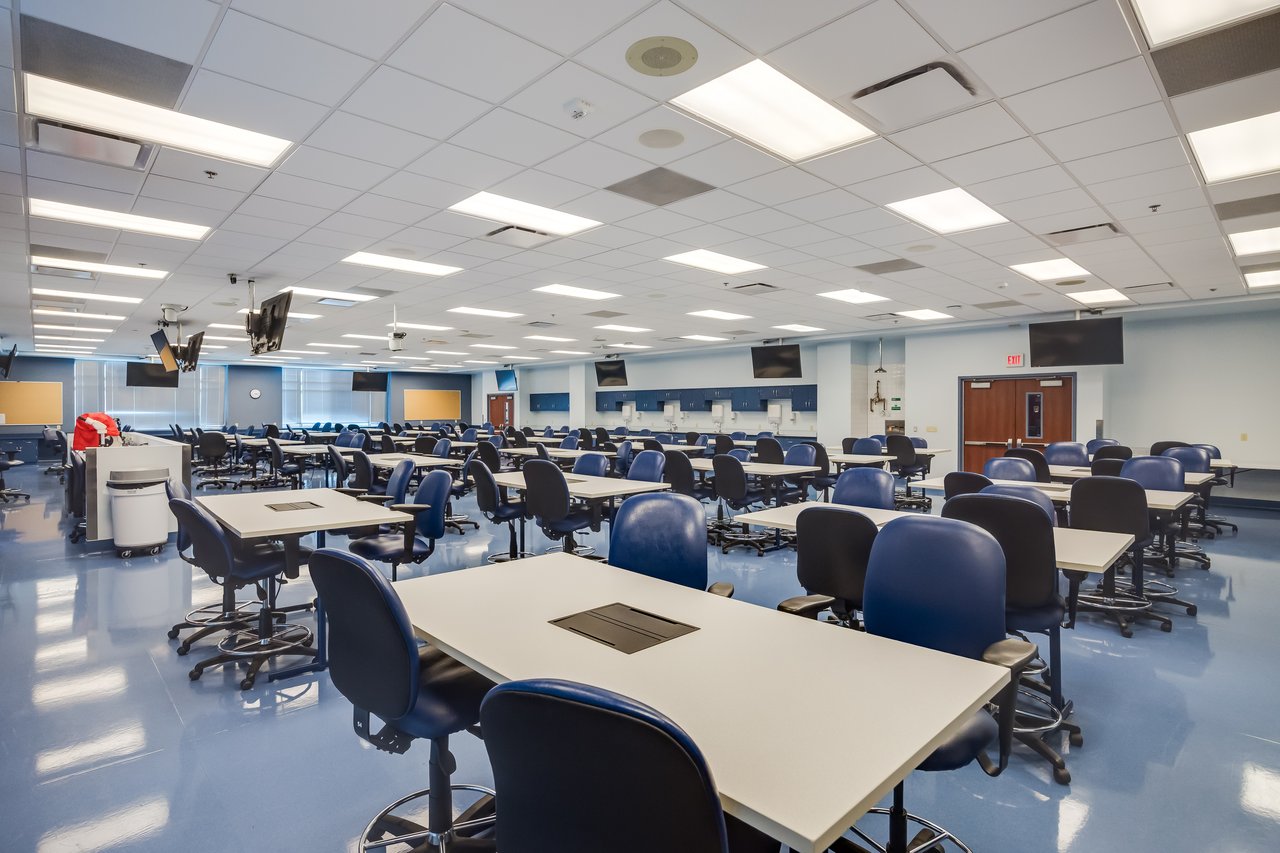 Spacious, modern classroom with rows of tables and blue chairs, featuring large screens and bright lighting.