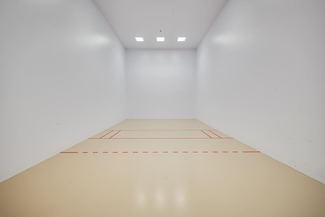 A minimal indoor racquetball court with beige flooring and red markings, surrounded by smooth white walls.