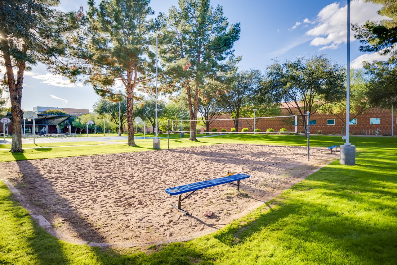 A sandy volleyball court surrounded by trees and grass, with a blue bench nearby and a clear sky above.