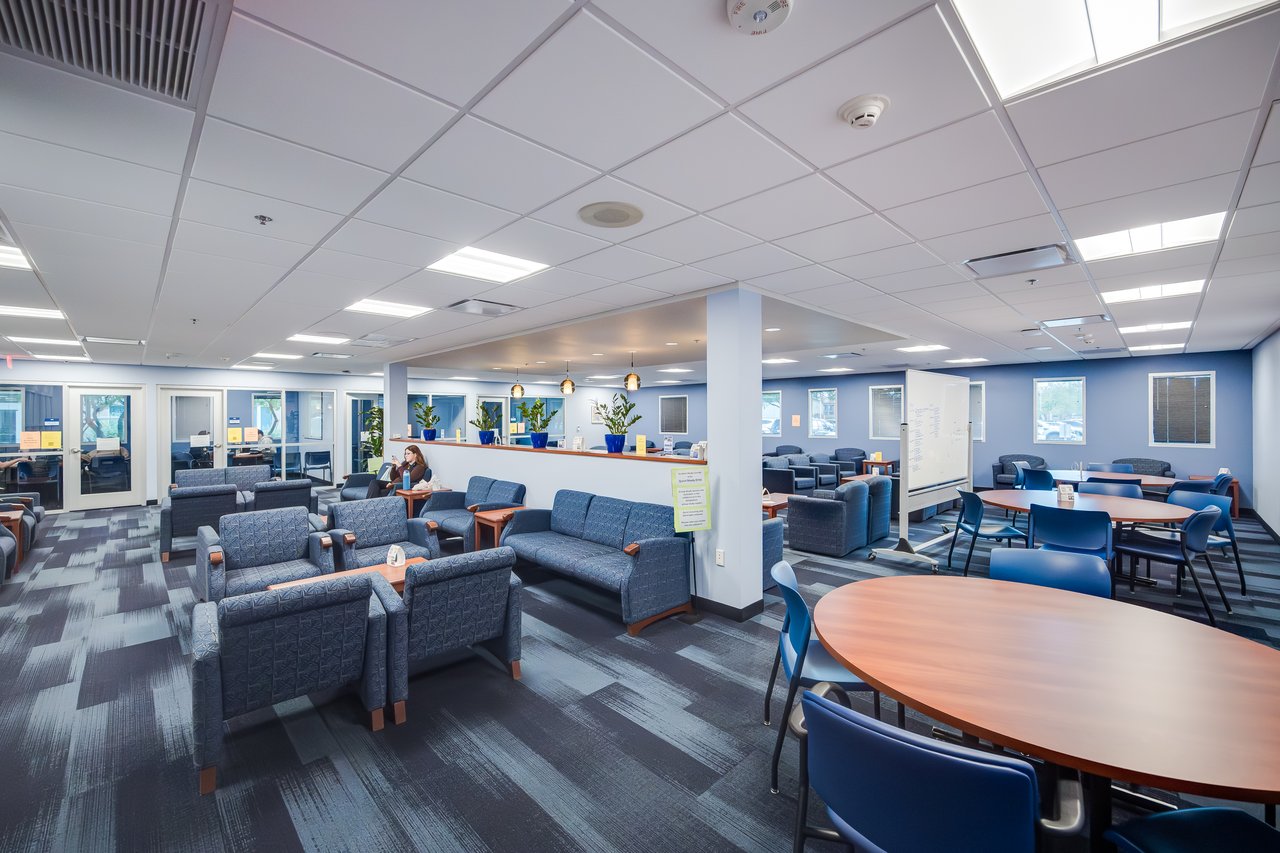 A spacious lounge with blue seating, circular tables, and plants, featuring large windows and a bright, open atmosphere.