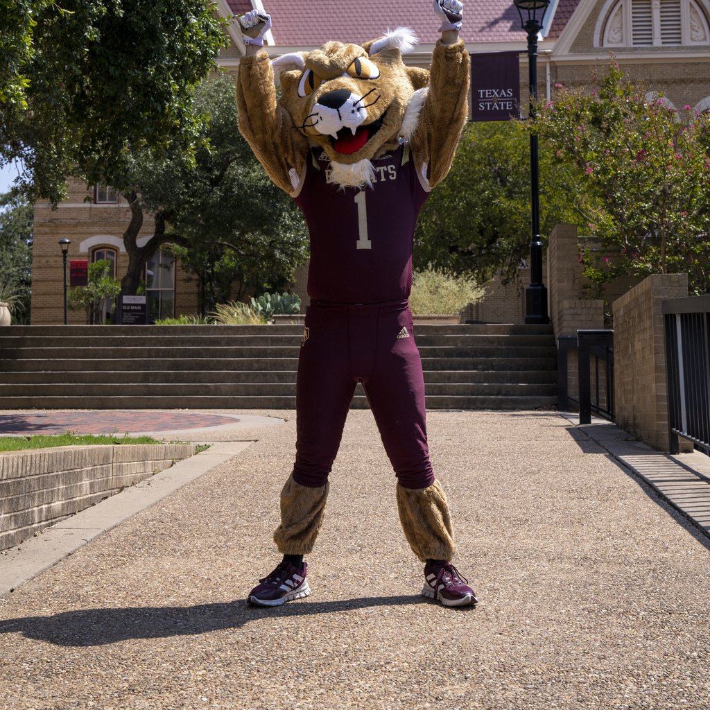 A costumed mascot with feline features strikes a triumphant pose in front of a university building.