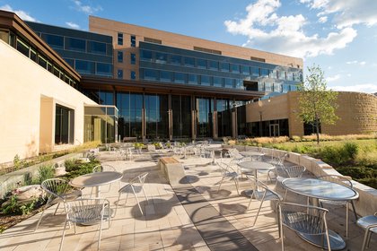 Modern building with glass exterior, surrounded by a patio with metal tables and chairs under a blue sky.